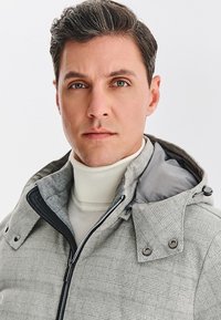 Szara kurtka w kratę z kapturem, wykonana z gładkiej tkaniny, z zamkiem błyskawicznym i czarnymi akcentami. Pod spodem biały golf.