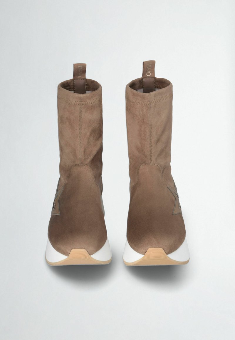 Bottines en daim marron avec un design ajusté, une semelle blanche et un détail de logo sur le côté. Texture douce et style minimaliste.