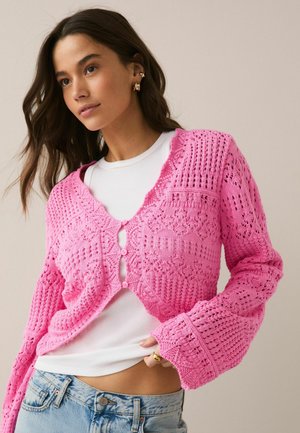 Junge Frau mit langen Haaren, die goldene Ohrringe trägt, eine rosa gestrickte kurze Strickjacke über einem weißen Top und hellblaue Jeans vor einem einfarbigen Hintergrund.