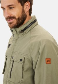 Olive green jacket with a high collar, zip and snap button closure, multiple front pockets, and a leather logo patch on the sleeve.