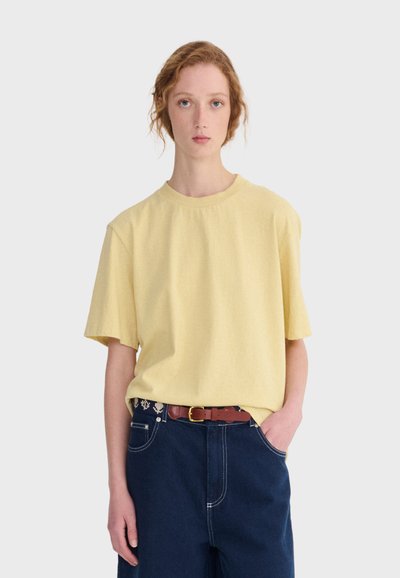 Personne à la peau claire et aux cheveux roux portant une chemise ample à manches courtes jaune pâle rentrée dans un pantalon bleu foncé avec une ceinture marron, main dans la poche.