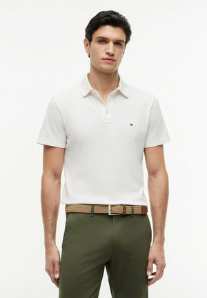 Mann trägt ein weißes kurzärmeliges Poloshirt, in olivgrüne Hose mit geflochtenem hellbraunem Gürtel gesteckt, steht vor einem einfarbigen Hintergrund.