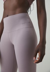 Leggings Nike a vita alta in morbido tessuto lilla, caratterizzati da una texture liscia, cuciture minime e un piccolo logo argentato sulla cintura.