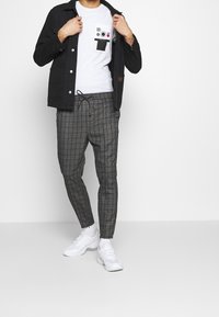 Svart jacka, vit grafisk t-shirt, grå rutiga byxor och vita sportskor. Outfiten har en avslappnad, lager-på-lager design.