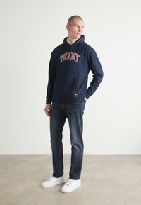 Námornícka mikina s kapucňou s červeným a bielym nápisom "TOMMY". Má predné vrecko, červené akcenty a je skombinovaná s tmavými džínsami a bielymi teniskami.