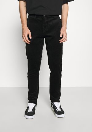 Mężczyzna w dopasowanych czarnych sztruksowych spodniach i czarno-białych trampkach na jednolitym jasnym tle.