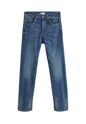 Ragazzo biondo in piedi con una mano in tasca, indossa una maglietta bianca, jeans blu e scarpe da ginnastica bianche su uno sfondo chiaro e semplice.