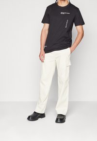 Svart kortärmad t-shirt med vita grafiska mönster, kombinerad med krämfärgade cargobyxor med sidofickor och en avslappnad passform, avslutad med svarta skor.