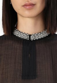 Blusa nera con texture plissettata e colletto gioiello adornato con piccole pietre di strass trasparenti e perline bianche. Chiusura a bottoni al collo.