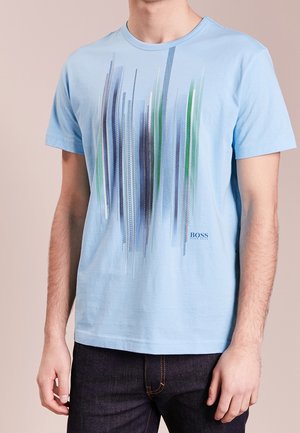 Mand iført en lyseblå T-shirt med korte ærmer og lodrette abstrakte striber i blå, grøn, hvid og marineblå, kombineret med mørke jeans.