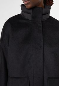 Persona che indossa un cappotto di lana nero con un alto colletto imbottito e tasche anteriori, mostrata dalle spalle fino a metà torace.