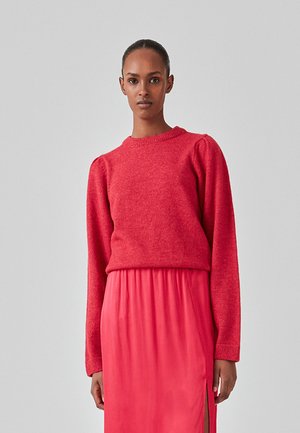 Rød strikket sweater med rund halsudskæring, lange ærmer og let puffede skuldre, båret over en lyserød satin-nederdel med sidespalter.