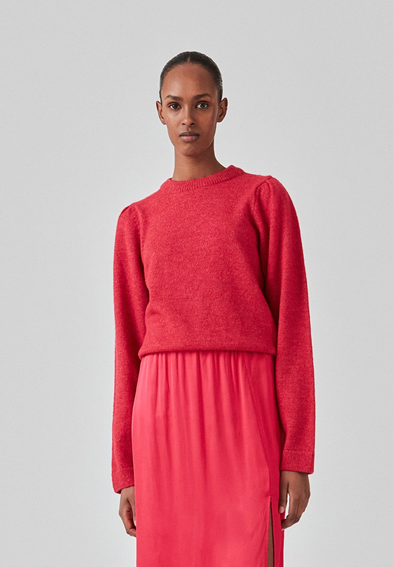 Rød strikket sweater med rund halsudskæring, lange ærmer og let puffede skuldre, båret over en lyserød satin-nederdel med sidespalter.
