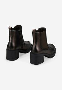 Par de botas de cuero negras hasta el tobillo con tacones gruesos, con cremallera lateral en una bota y panel elástico en la otra, sobre fondo blanco.