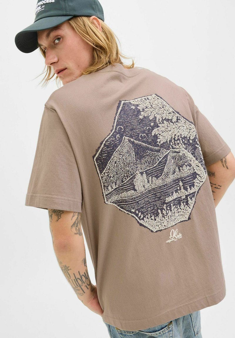 Brun bomulds t-shirt med et stort marineblåt grafisk tryk af bjerge og træer på ryggen, med korte ærmer og en afslappet pasform.