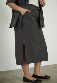 Femme portant une jupe midi foncée avec une fente devant, chemise assortie, top blanc et ballerines noires, debout sur un sol en béton.