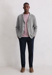 Grauer Wollcardigan über einem pinken Hemd, kombiniert mit navyblauen Hosen und braunen Wildlederschuhen. Der Cardigan verfügt über einen Kragen und zwei vordere Taschen.