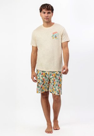 Hombre de pie descalzo, vestido con una camiseta crema con un pequeño gráfico de surf y pantalones cortos estampados con tablas de surf y palmeras.