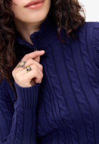 Navy blue cable-knit sweater with a high collar and zip detail. Features ribbed cuffs and textured patterns. Accessories include two rings.