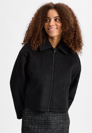 Zwarte, ritsjack met een kraag, gemaakt van zacht, textuurachtig materiaal. Beschikt over twee zijzakken en een cropped silhouet.