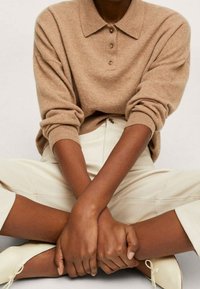 Osoba siedząca w pozycji skrzyżowanej, ubrana w beżowy sweter zapinany na guziki, jasne spodnie i dopasowane baleriny, z dłońmi złożonymi na kolanach.