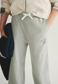 Grijze sweatpants met een trekkoord in de taille, zijzakken en blauwe geborduurde tekst met de opdruk "New York S. 1986 Athletics." Zacht, gestructureerd materiaal.
