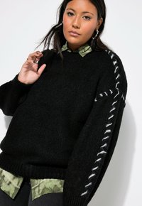 Schwarzer Strickpullover mit strukturierter Oberfläche und weißen Nähdetails an einem Ärmel. Über einem grün gemusterten Hemd getragen.