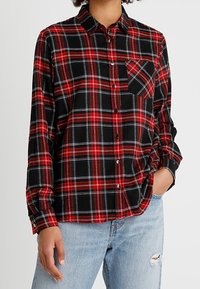 Camicia in flanella a quadri rossi e neri con maniche lunghe, chiusura con bottoni e una tasca sul petto. Texture morbida e vestibilità comoda.