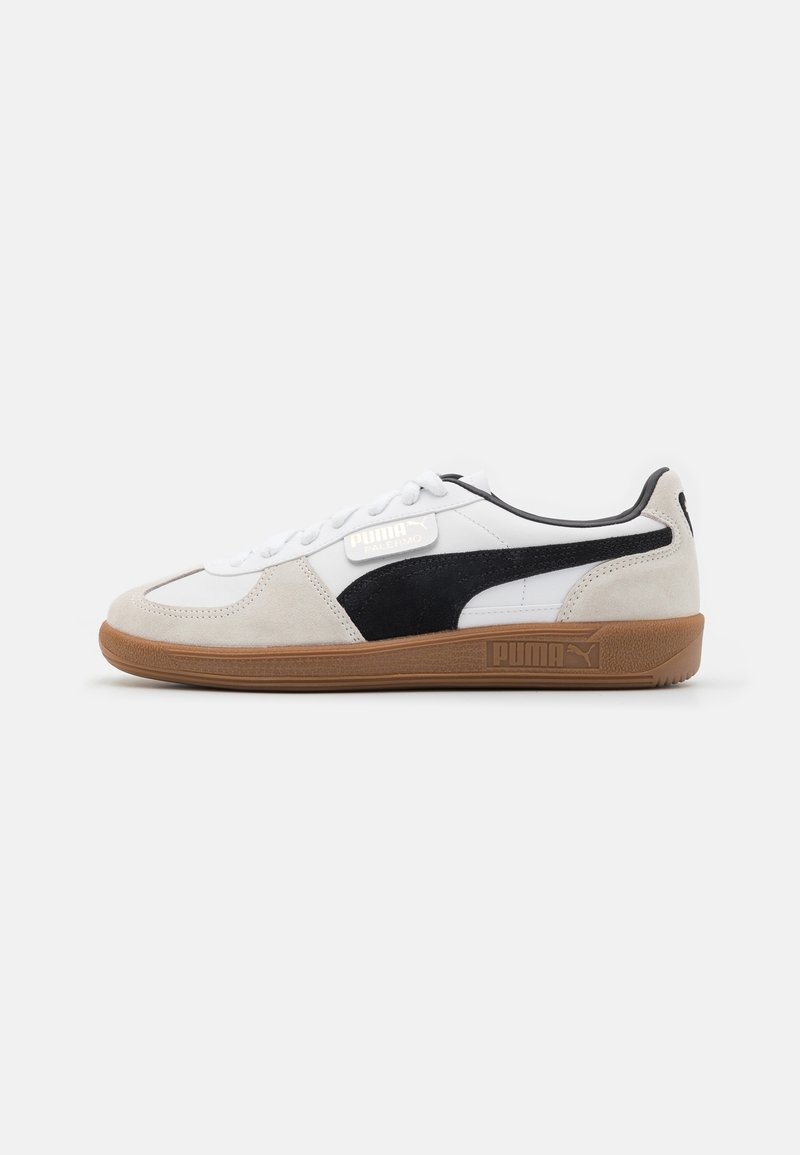 Basket basse Puma blanche avec des accents en daim beige, bande latérale noire, lacets blancs et semelle en caoutchouc marron sur un fond neutre.