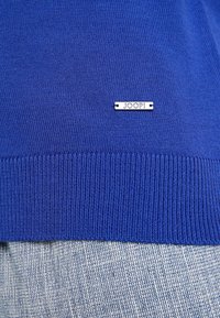 Blauer Strickpullover mit geripptem Saum, ausgestattet mit einem silbernen Metall-Logoetikett mit der Beschriftung "JOOP!" in der unteren linken Ecke. Weiche Textur über das gesamte Oberteil.