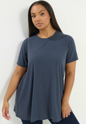 Mujer con cabello largo y oscuro, vestida con una blusa azul oscuro de manga corta, holgada y acanalada, de pie contra una pared blanca.