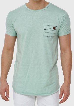 Homme portant un t-shirt à manches courtes vert clair avec une poche rayée noire et un pantalon blanc, devant un fond uni.
