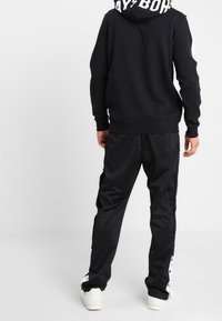 Svart hoodie med vit text på huvan, kombinerad med svarta joggerbyxor som har sidodetaljer och vit kant.