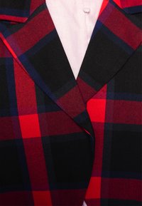 Gros plan sur le revers d'une veste à carreaux rouge, noire et bleue sur une chemise blanche à boutons.