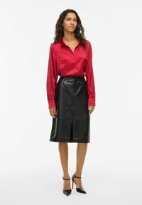 Woman standing, wearing a red satin button-up blouse, black leather knee-length skirt with front slit, and black high-heeled shoes.
