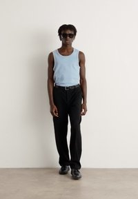 Débardeur sans manches bleu clair, pantalon noir et chaussures noires. Design ajusté avec encolure standard. Le mannequin se tient contre un mur neutre.