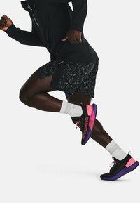 Löpare i svarta sportshorts, svarta och rosa joggingskor, vita strumpor och en svart långärmad tröja fångad mitt i steget.