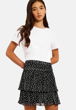 Jonge vrouw met lang haar die een eenvoudig wit T-shirt met korte mouwen draagt en een zwarte gelaagde rok met witte stippen.