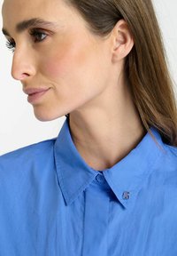Camisa de color azul claro con cuello y una textura suave, que presenta un pequeño logo metálico en el cuello y un cierre de botones en la parte delantera.