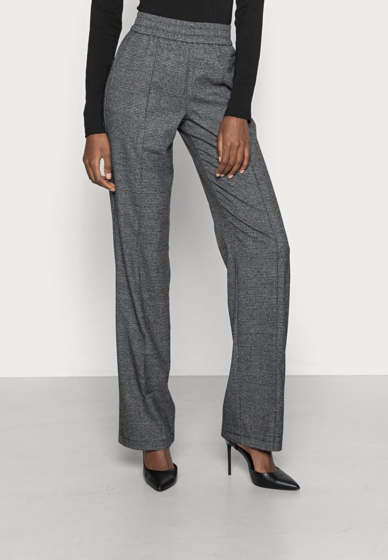 Femme portant un pantalon large à carreaux gris et des talons hauts noirs à bout pointu, debout devant un fond uni clair.