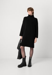 Zwarte geribbelde gebreide jurk met een hoge col, lange mouwen en een losse fit, gecombineerd met zwarte enkellaarsjes en een zwarte tas.