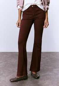 Persona con pantalones de campana burdeos con bolsillos delanteros, combinados con zapatos planos negros con tachuelas, de pie sobre un suelo gris frente a una pared clara.