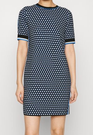 Femme portant une robe noire à manches courtes avec un motif à pois bleu et blanc et une bordure bleue aux manches et à l'encolure.
