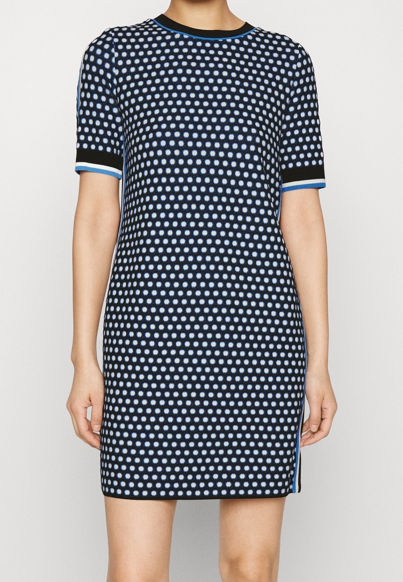 Femme portant une robe noire à manches courtes avec un motif à pois bleu et blanc et une bordure bleue aux manches et à l'encolure.