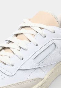 Beli usnjeni tenisici z bež dodatki, ki ima teksturirane pregrade, ravne bele vezalke in subtilen okno detajl na strani.