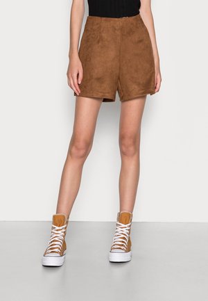 Person trägt braune Wildleder-Shorts, schwarzes Oberteil und beige High-Top-Plateau-Sneaker mit weißen Schnürsenkeln, steht vor einem einfarbigen Hintergrund.