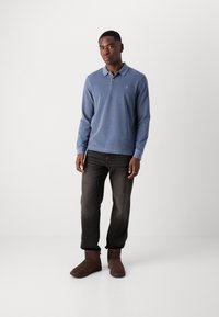 Polo shirt bleu à manches longues avec col, associé à un jean foncé et des bottes chelsea en suède marron. Présente un petit logo sur la poitrine.