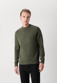Groene trui van een gestructureerde stof, met een geribbelde ronde hals en zoom, lange mouwen en een klassieke pasvorm. Zwarte broek wordt gedragen.