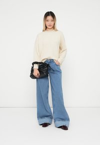 Maglione lavorato a maglia crema, jeans a gamba larga azzurri chiari con ampi risvolti, borsa a tracolla nera con lacci, e scarpe scure con punta affusolata.
