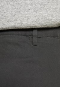 Detailní záběr na pas šedých kalhot s poutkem na opasek a světlým šedým pleteným svrškem nad ním.